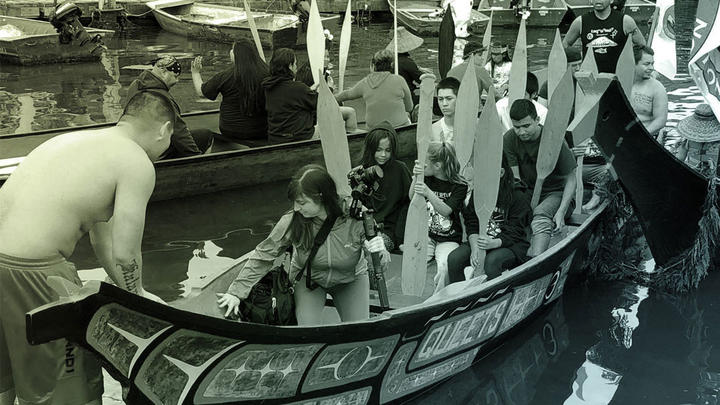 A man steadies a canoe that reads QUEETS on the side, which contains Crosscut video producer who is holding a camera on a tripod, and children and adults holding paddles